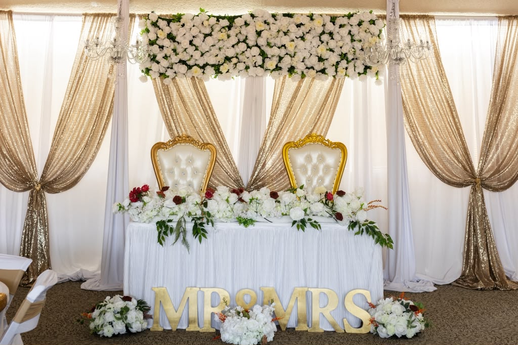 A head table with gold throne chairs, white florals and gold MR & MRS letters in front of gold sequin drapes